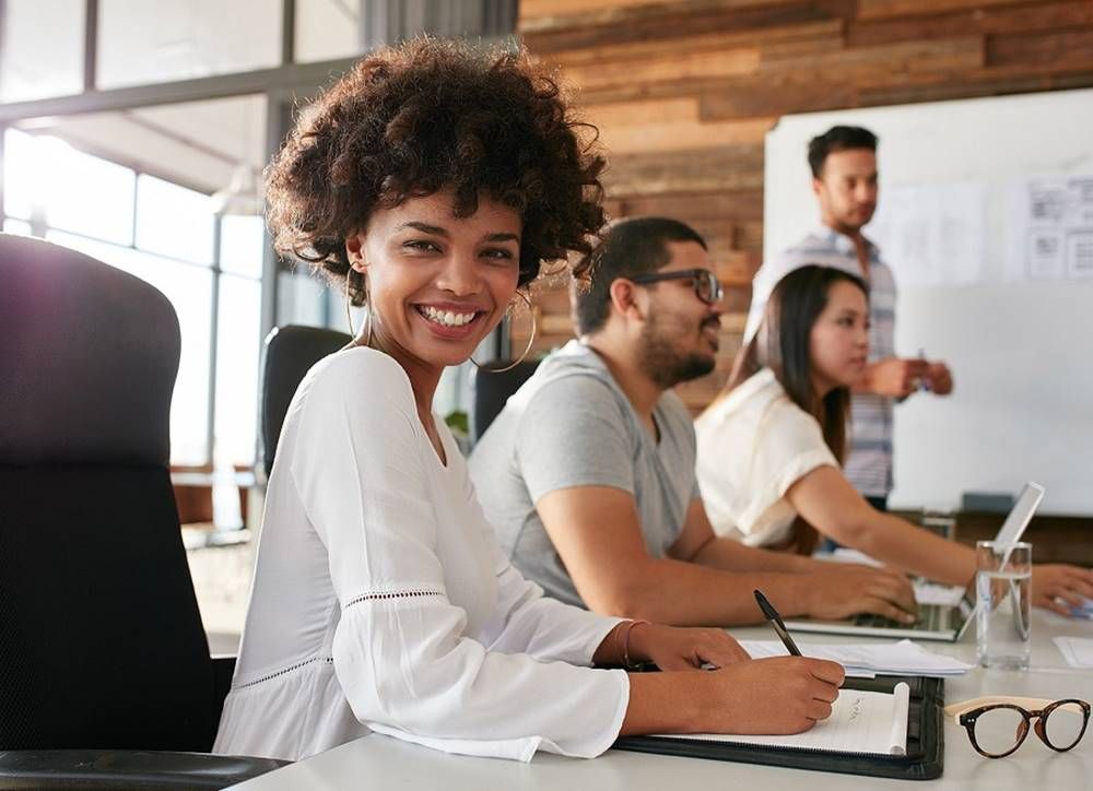 Vrouw lacht in camera en zit achter bureau.